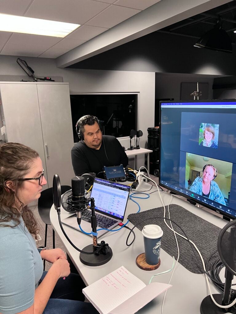People in a podcast recording studio, with a video call on screen. Transcribed Text: The notebook has handwritten text, partially visible, in red ink.