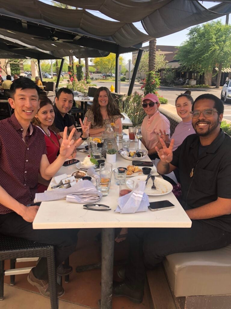 A group of seven people sitting around a table at an outdoor restaurant, smiling and making peace signs.